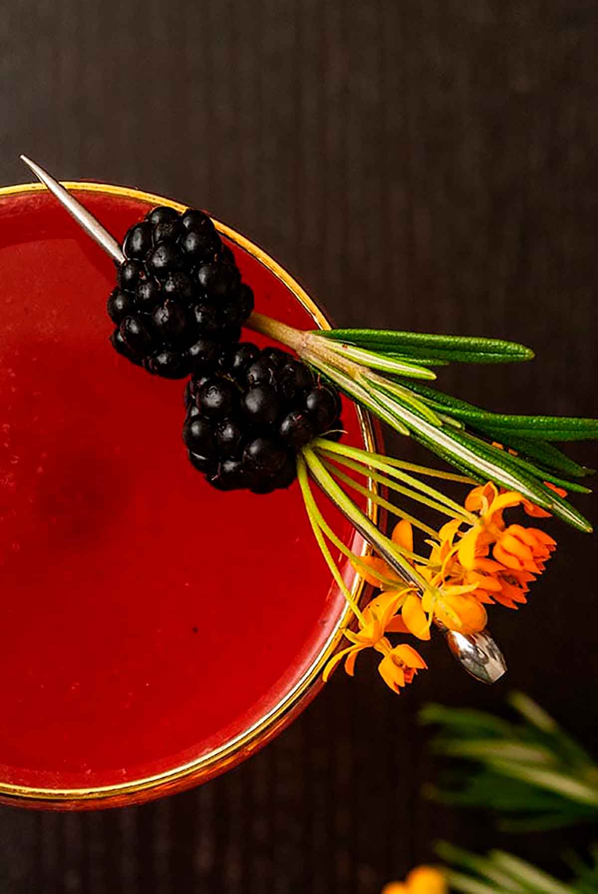 The top of a cocktail on a wood table, garnished with flowers and blackberries.