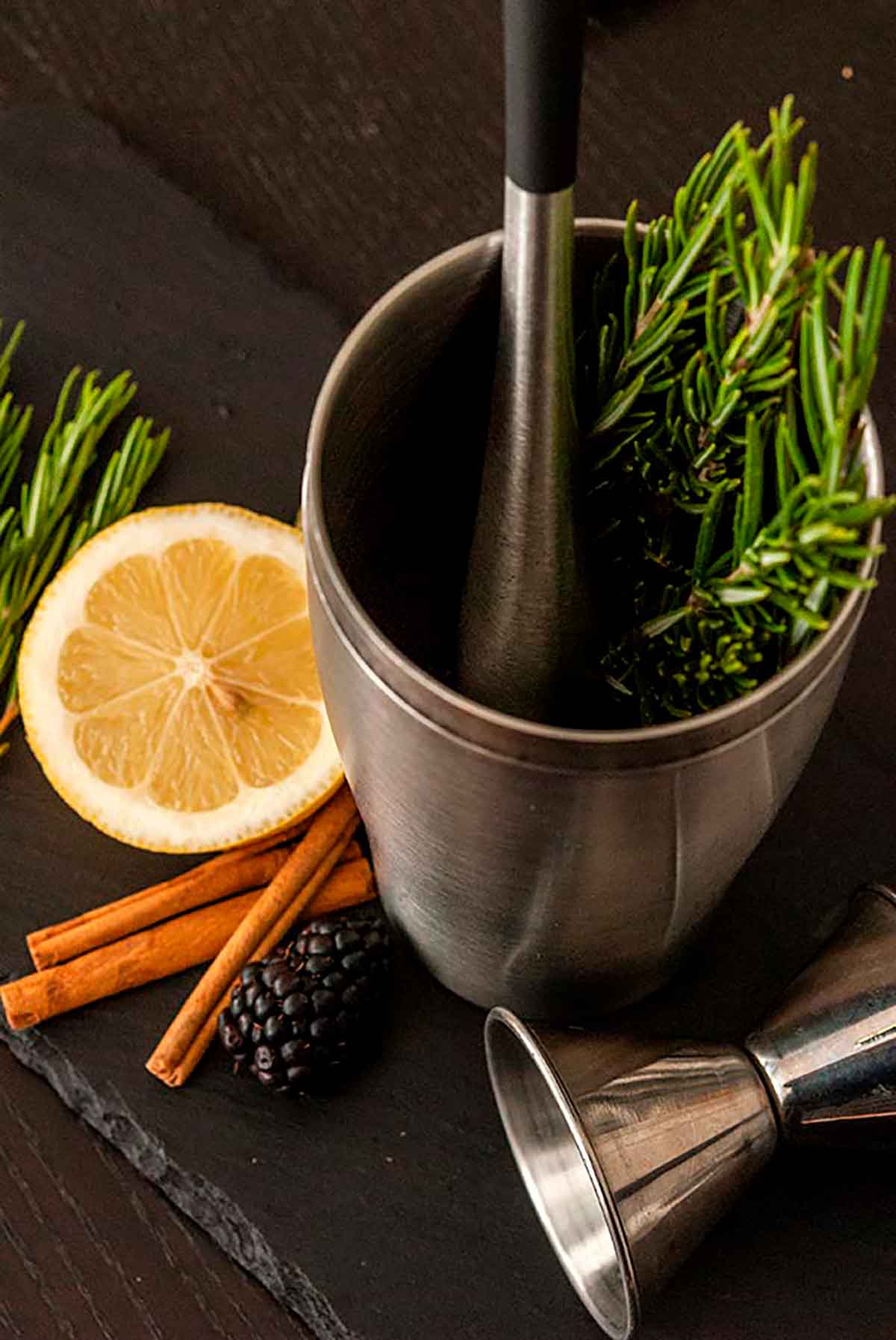 A cocktail shaker with rosemary inside, surrounded by cinnamon sticks, a lemon slice, blackberries and a cocktail jigger.