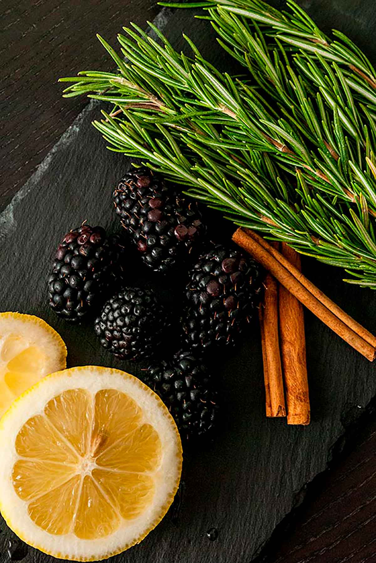 Sliced lemons, 5 blackberries, 2 cinnamon sticks and a few sprigs on rosemary on a dark slate.