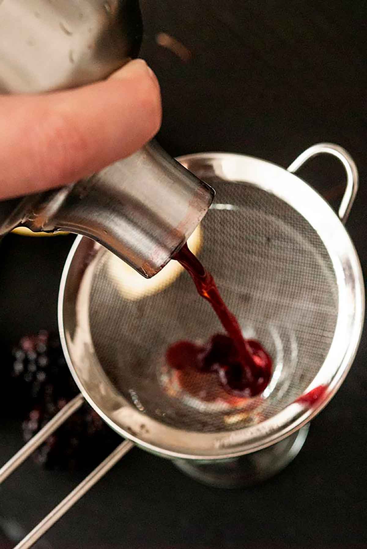 A cocktail shaker pouring blackberry mixture into a strainer above a glass.