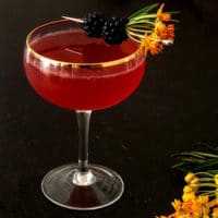 A cocktail on a wood table, garnished with flowers and blackberries with a few flowers on the table.