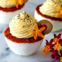 3 deviled eggs with a spiced rim on a marble plate, garnished with a flower and olive. with flowers on the side of the plate.