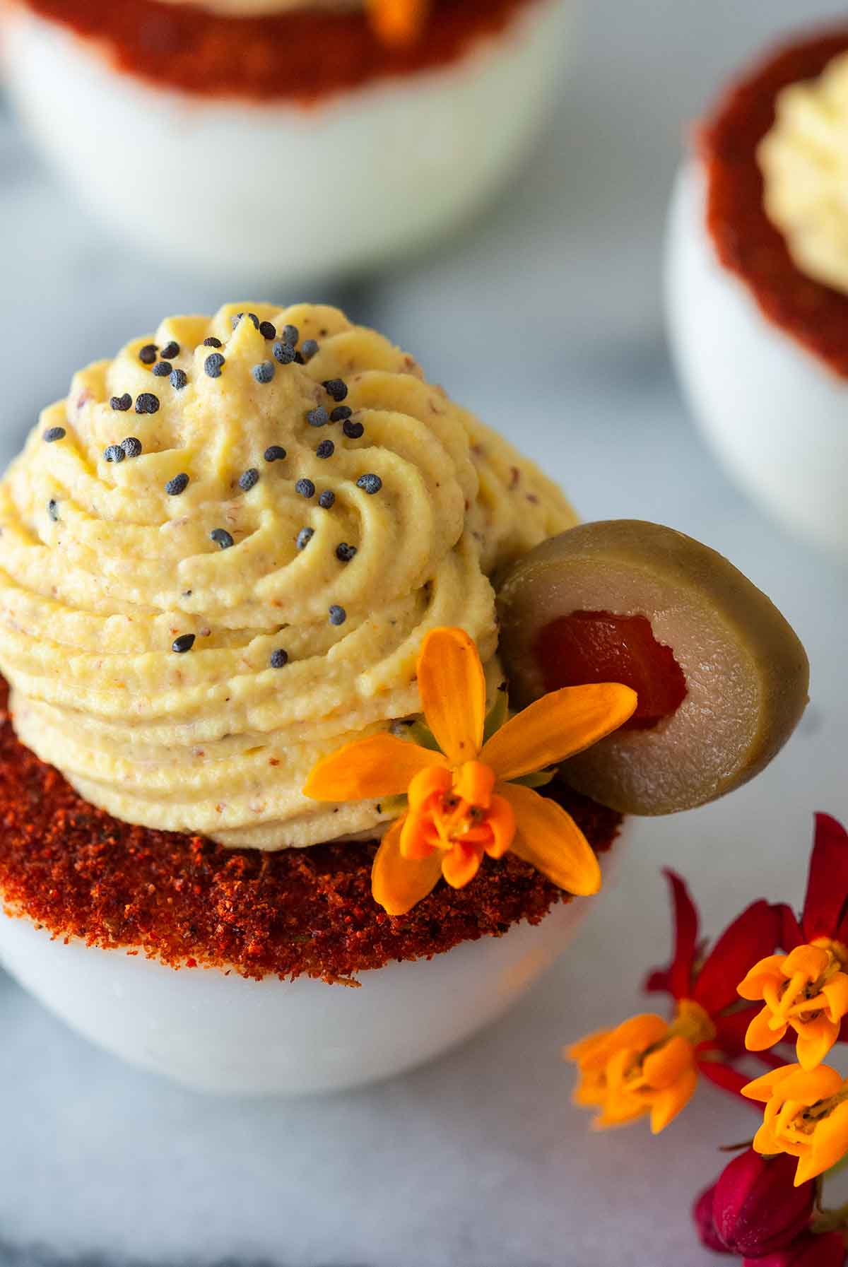 A deviled egg with a spiced rim on a marble plate, garnished with a flower and olive.