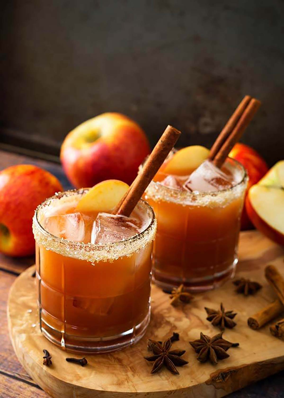 2 cocktails garnished with cinnamon sticks, apple slices and sugar rims, surrounded by a few apples and spices on the base.