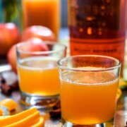 2 cocktails on a tray in front of a bottle of bourbon, surrounded by orange slices and apples.