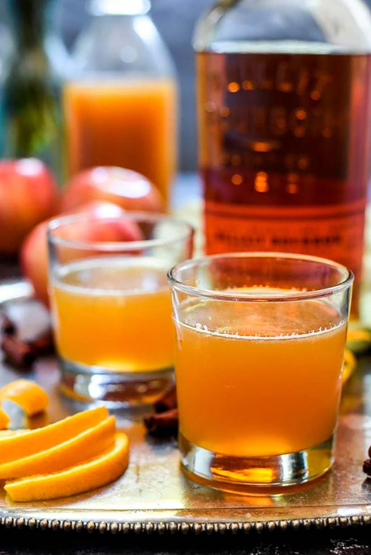 2 cocktails on a tray in front of a bottle of bourbon, surrounded by orange slices and apples.