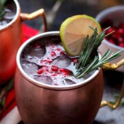 A cocktail in a mug, garnished with rosemary, a lemon and pomegranate seeds beside another cocktail and bowl of more seeds.