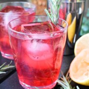 A cranberry cocktail garnished with rosemary beside 2 lemons and another cocktail in the background.