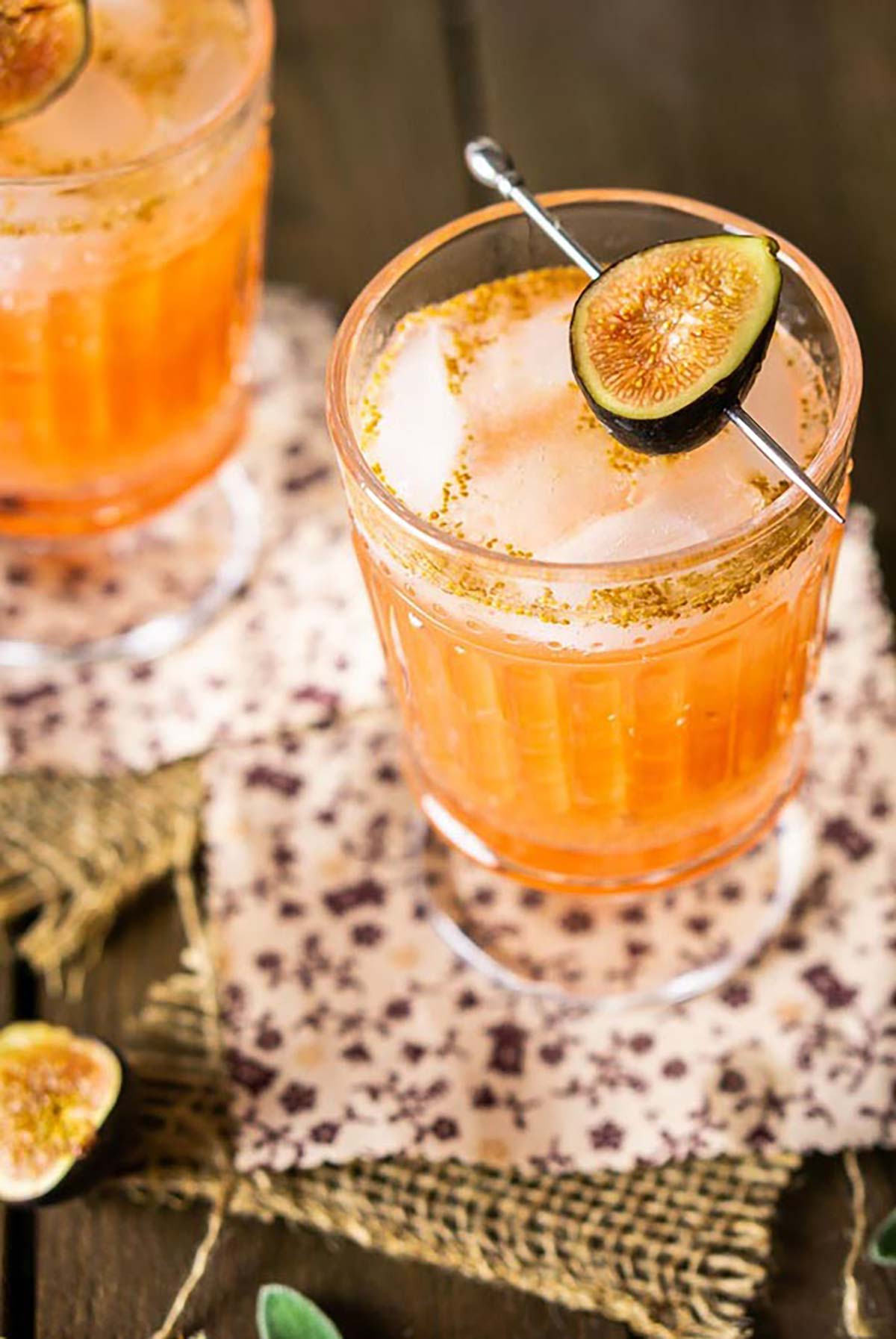 2 cocktails garnished with figs on a table with burlap and floral fabric coasters.