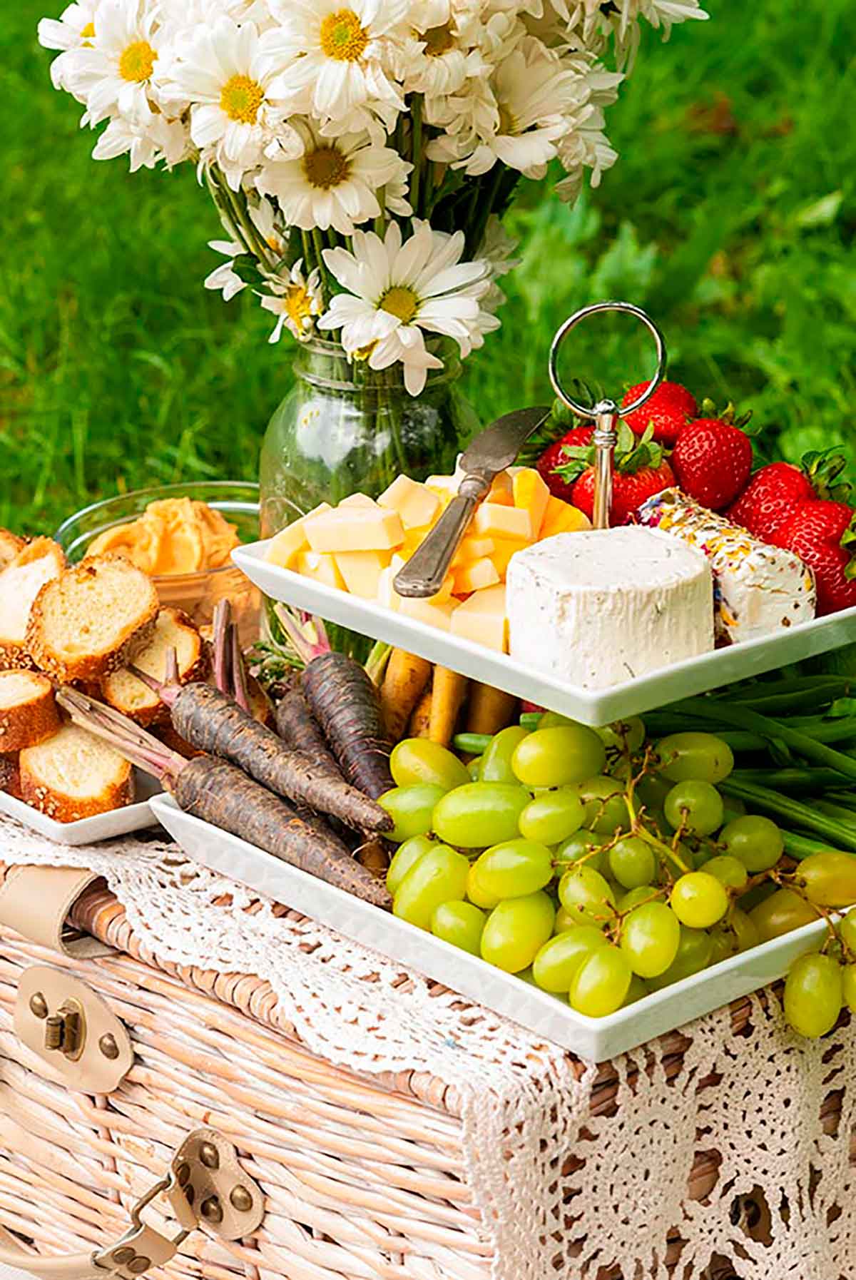 A 2 tiered plate with fruits, vegetables, cheese and bread, placed on a picnic basket, beside a jar of daisies on grass.