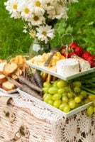 A 2 tiered plate with fruits, vegetables, cheese and bread, placed on a picnic basket, beside a jar of daisies on grass.