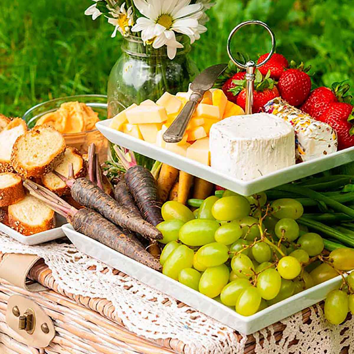 A 2 tiered plate with fruits, vegetables, cheese and bread, placed on a picnic basket, beside a jar of daisies on grass.