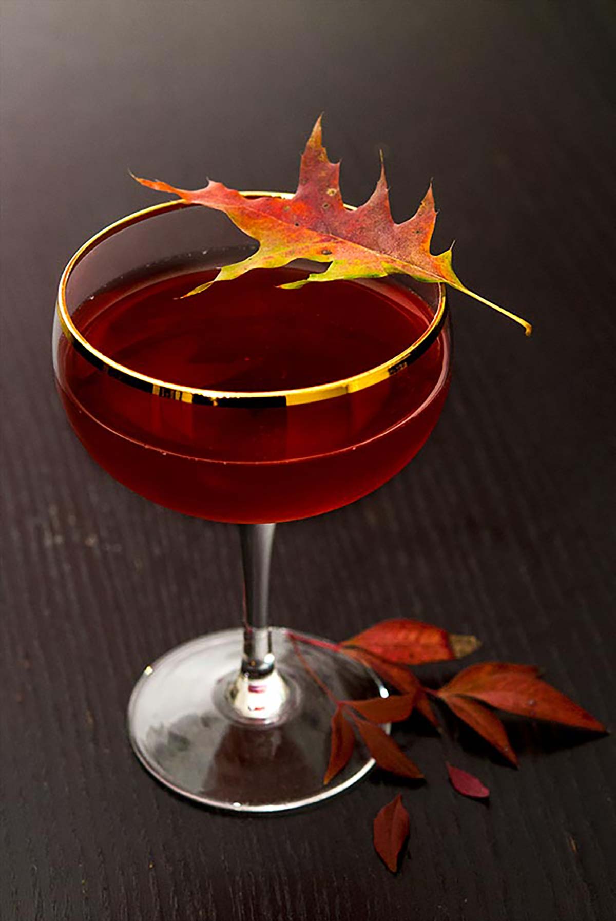 A cocktail garnished with a colorful, autumn leaf on a dark wood table with leaves at its base.