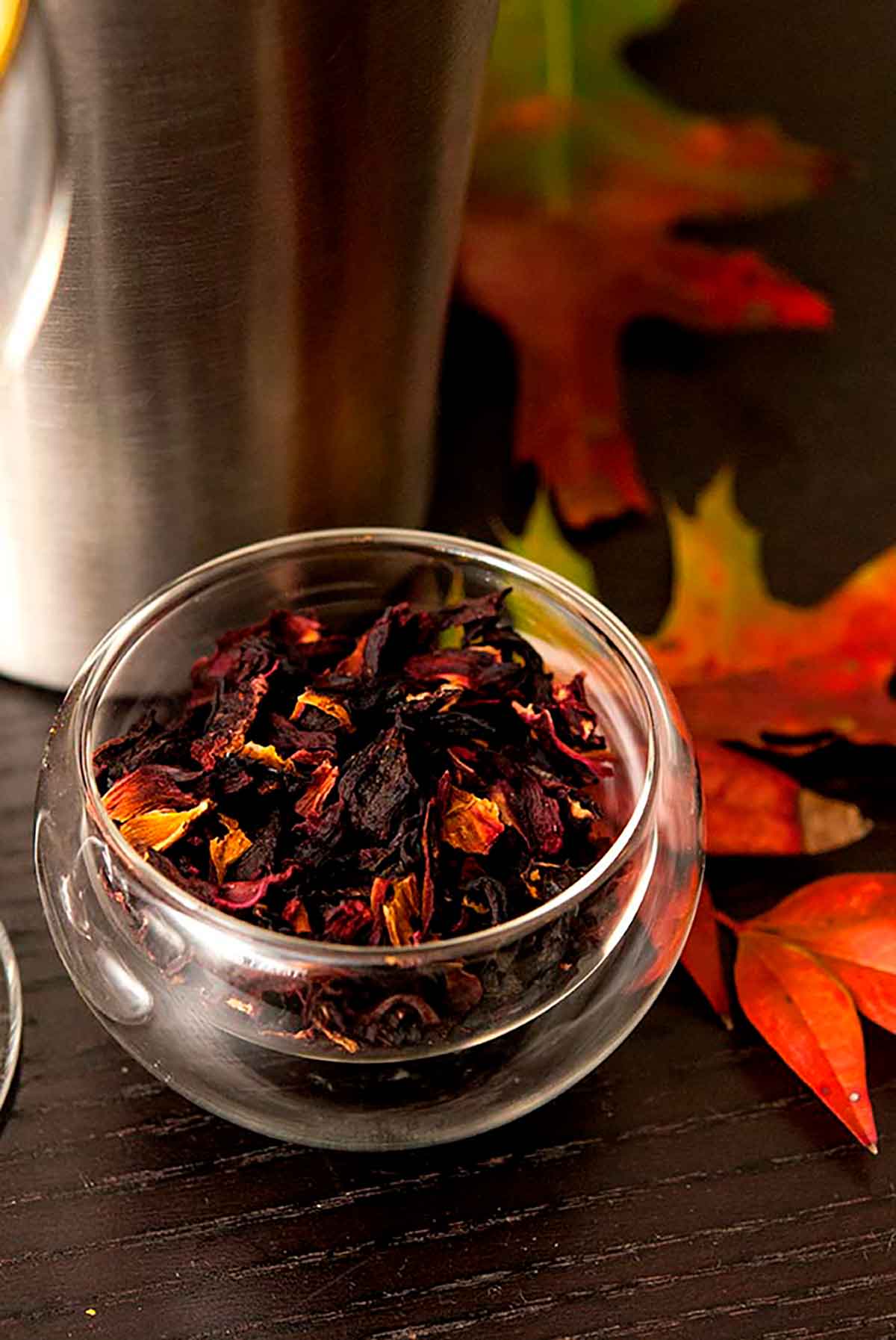 A small bowl of hibiscus flowers on a table with a few autumn leaves, in front of a cocktail shaker.