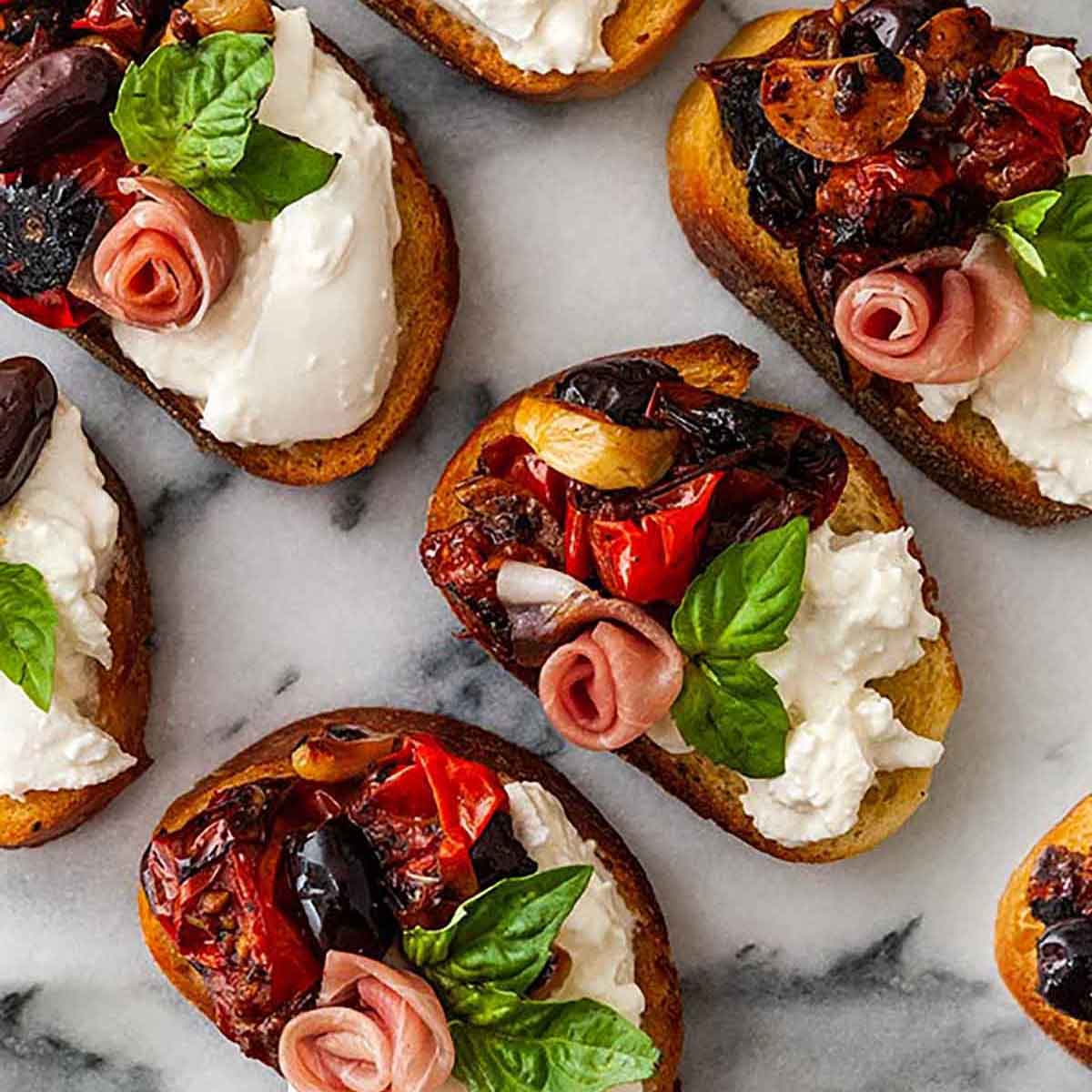 7 bruschetta on a marble slate with tomato, burrata and a prosciutto rose, garnished with basil.