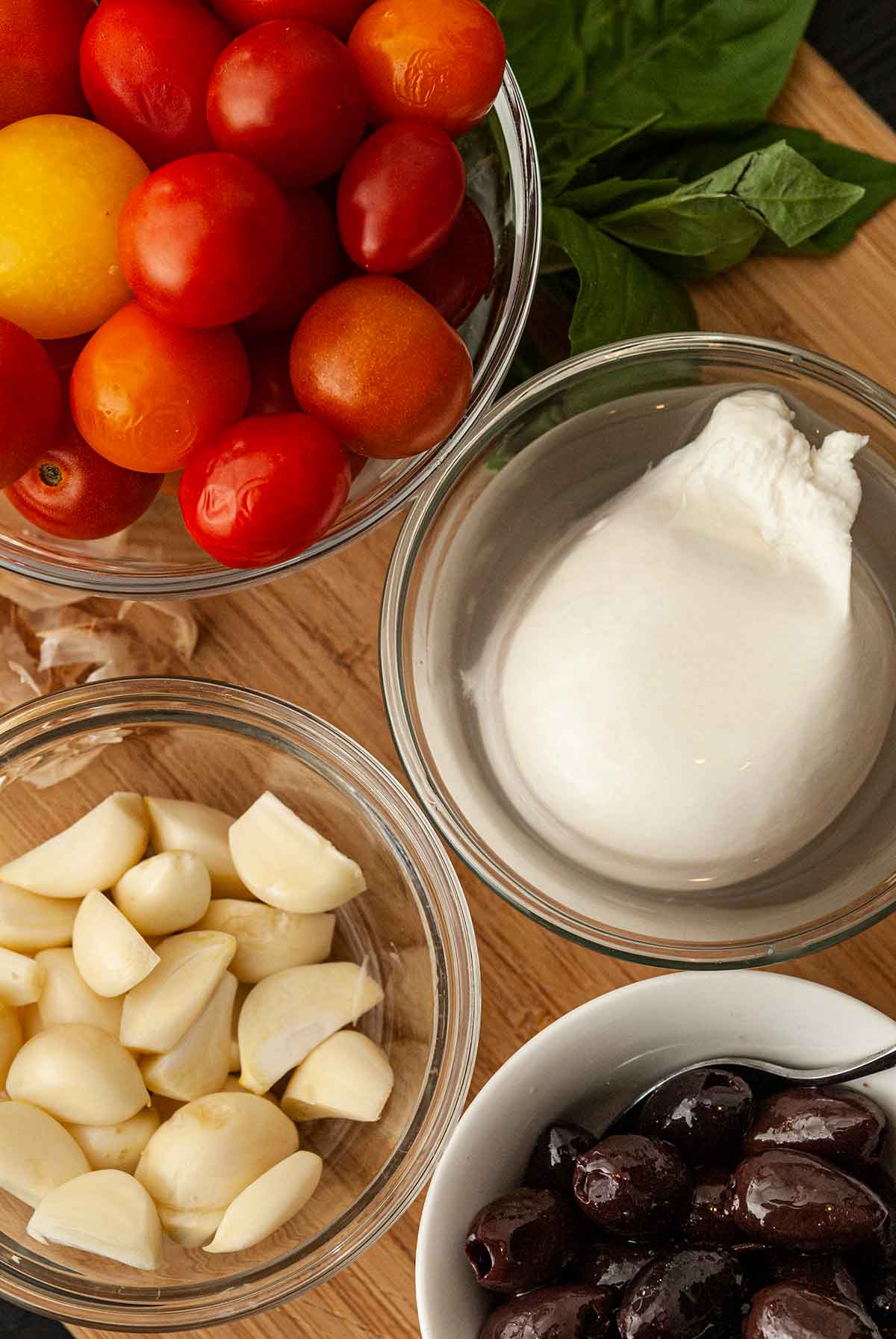 Small bowls on a wooden board with mozzarella, tomatoes, olives and garlic.