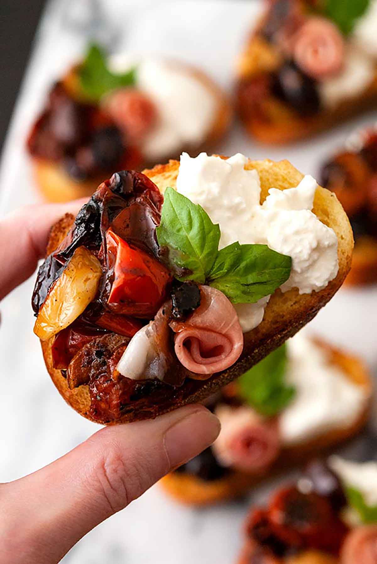 Fingers holding a bruschetta with tomato, burrata and a prosciutto rose, garnished with basil.