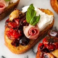 A bruschetta with tomato, burrata and a prosciutto rose, garnished with basil on a marble plate.