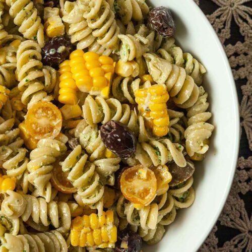 A bowl of pasta salad with fresh corn, tomatoes and olives on a lace tablecloth.