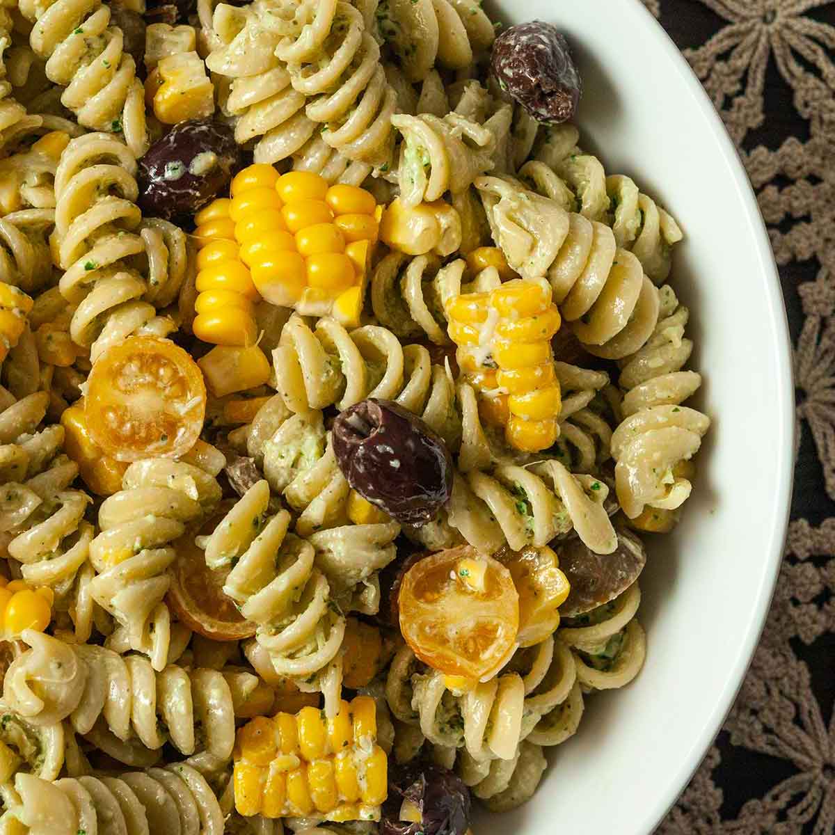 A bowl of pasta salad with fresh corn, tomatoes and olives on a lace tablecloth.
