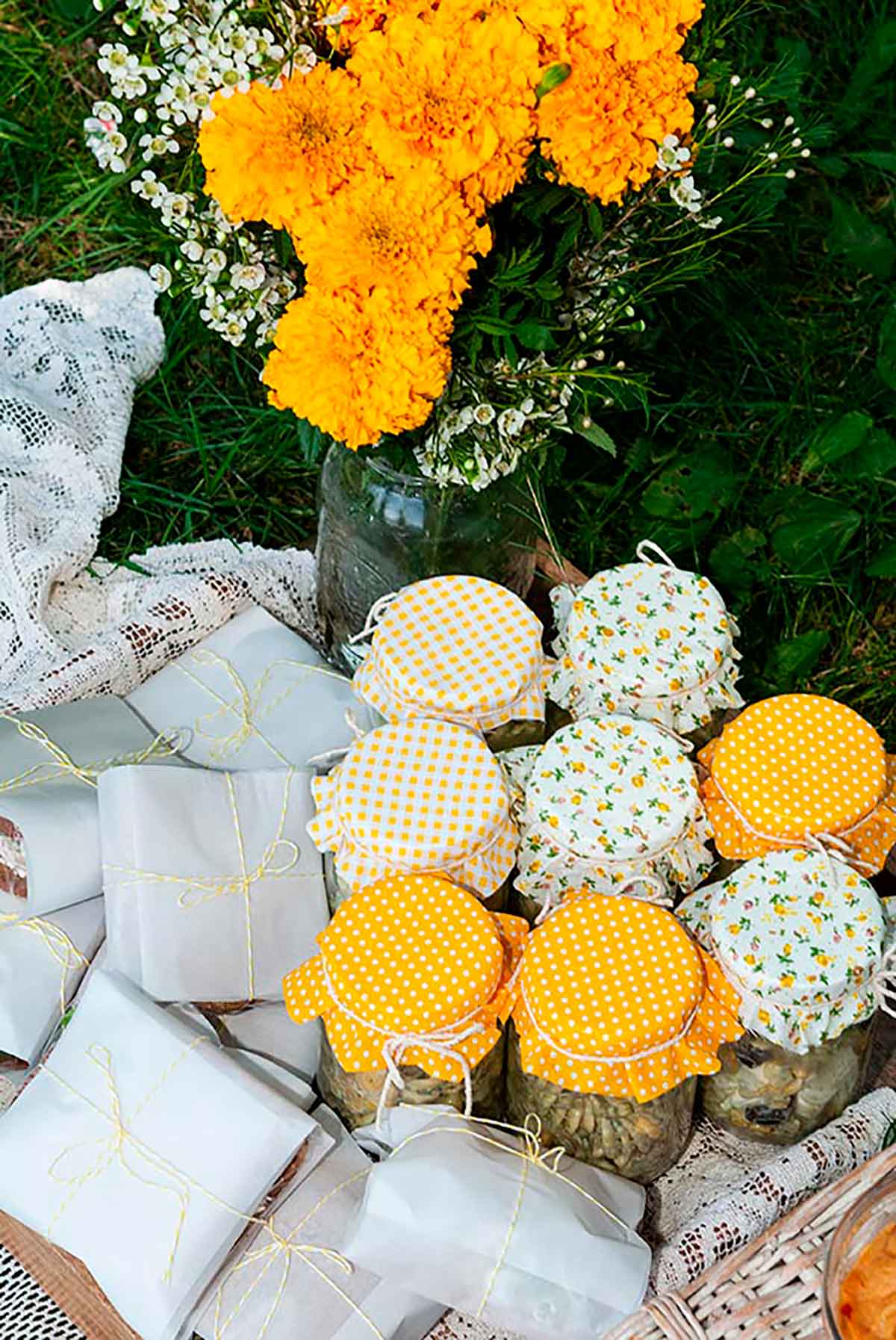 Fabric-covered jars of pasta salad and wrapped sandwiches in a lace-lined tray with a bouquet of marigold flowers.