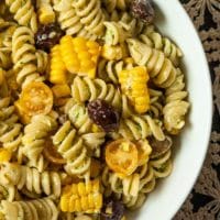 A bowl of pesto pasta salad with corn, olives, cherry tomatoes and mozzarella.