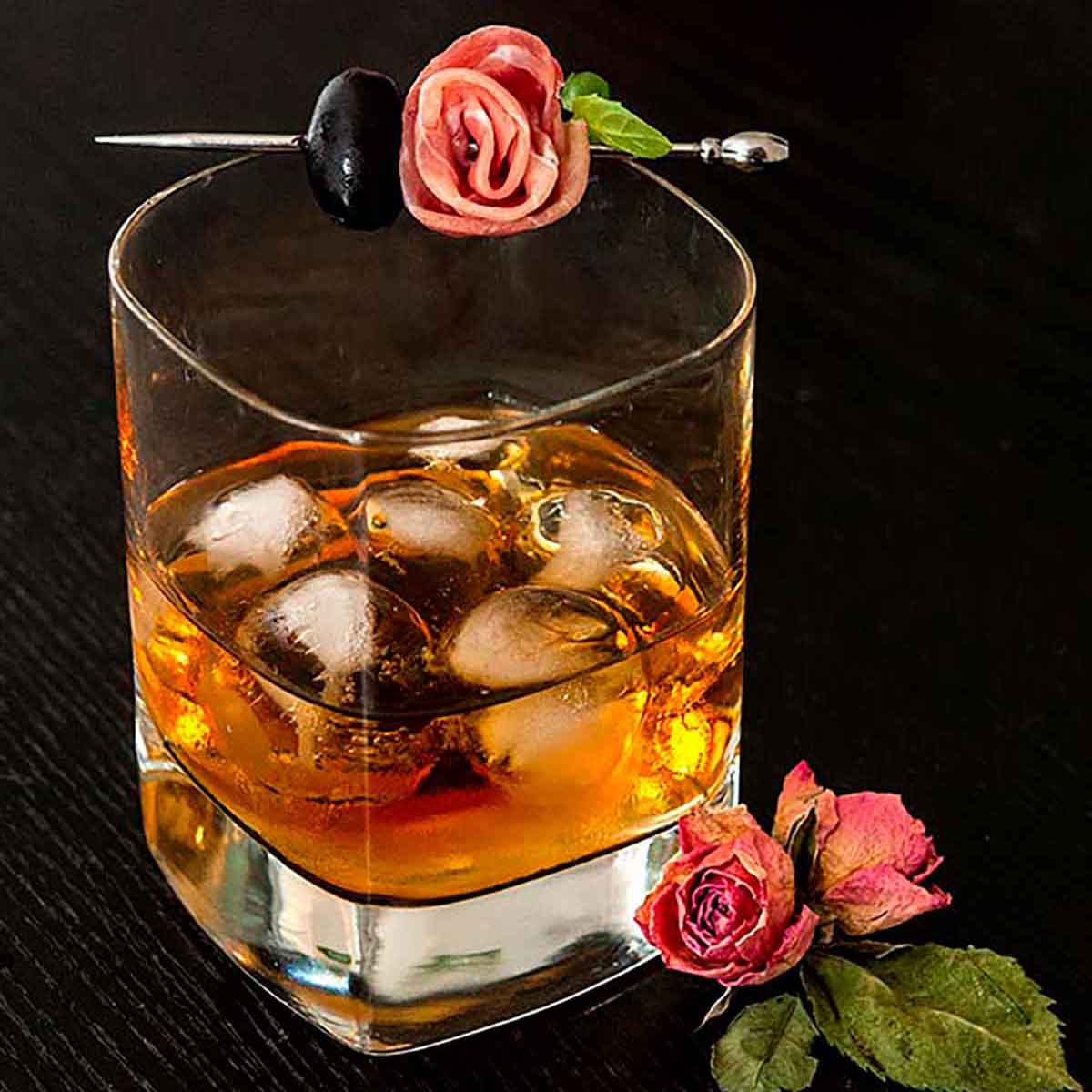 A whisky cocktail on a black table, surrounded by pink roses, garnished with a prosciutto rose and olive.
