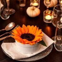 A sunflower in a soup bowl on a set dinner table, decorated with tea lights and small, white pumpkins.