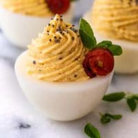 A deviled egg, garnished with a small bacon rose and a few tiny thyme leaves on a marble plate, in front of 2 other eggs.
