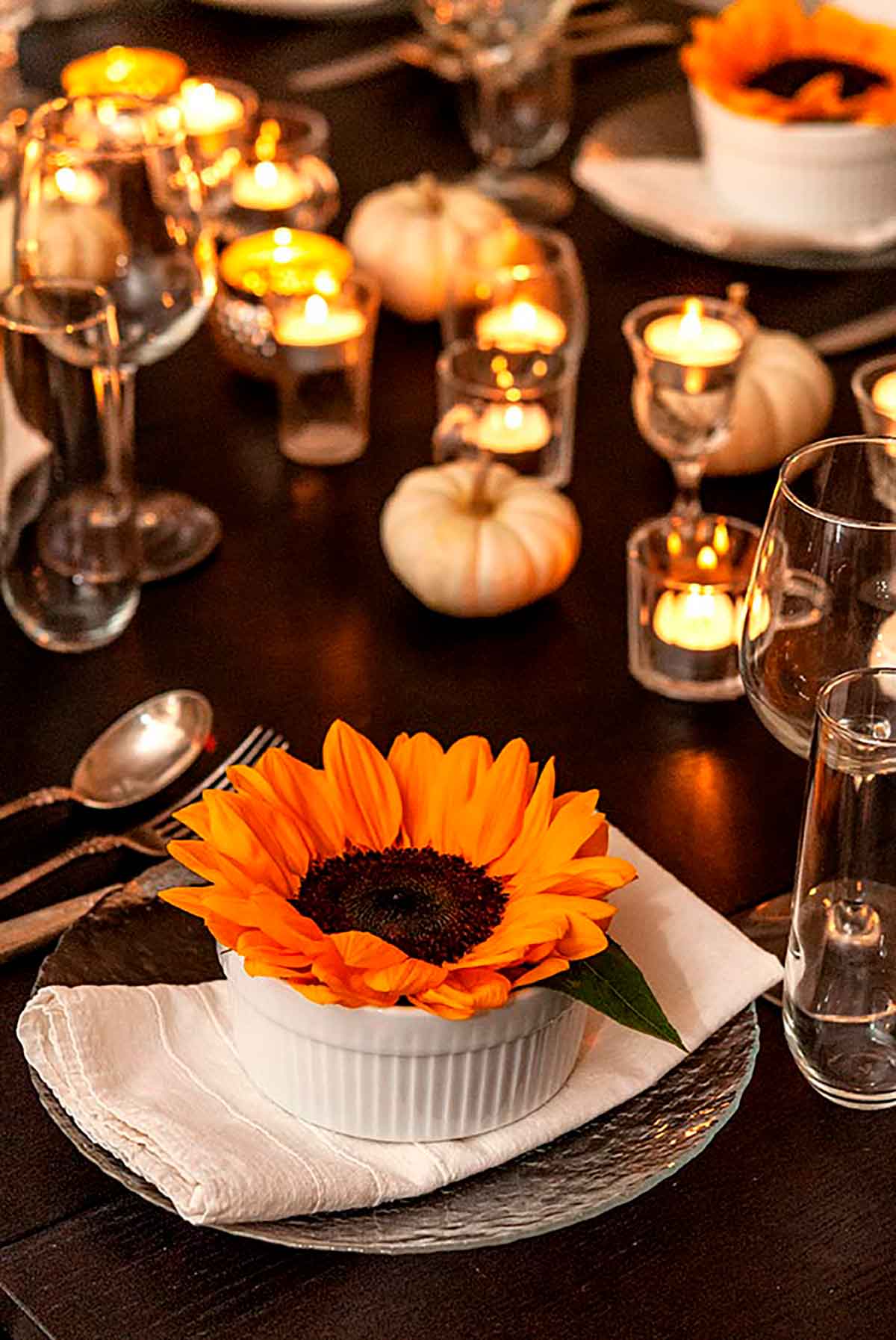 A sunflower in a soup bowl on a set dinner table, decorated with tea lights and small, white pumpkins.