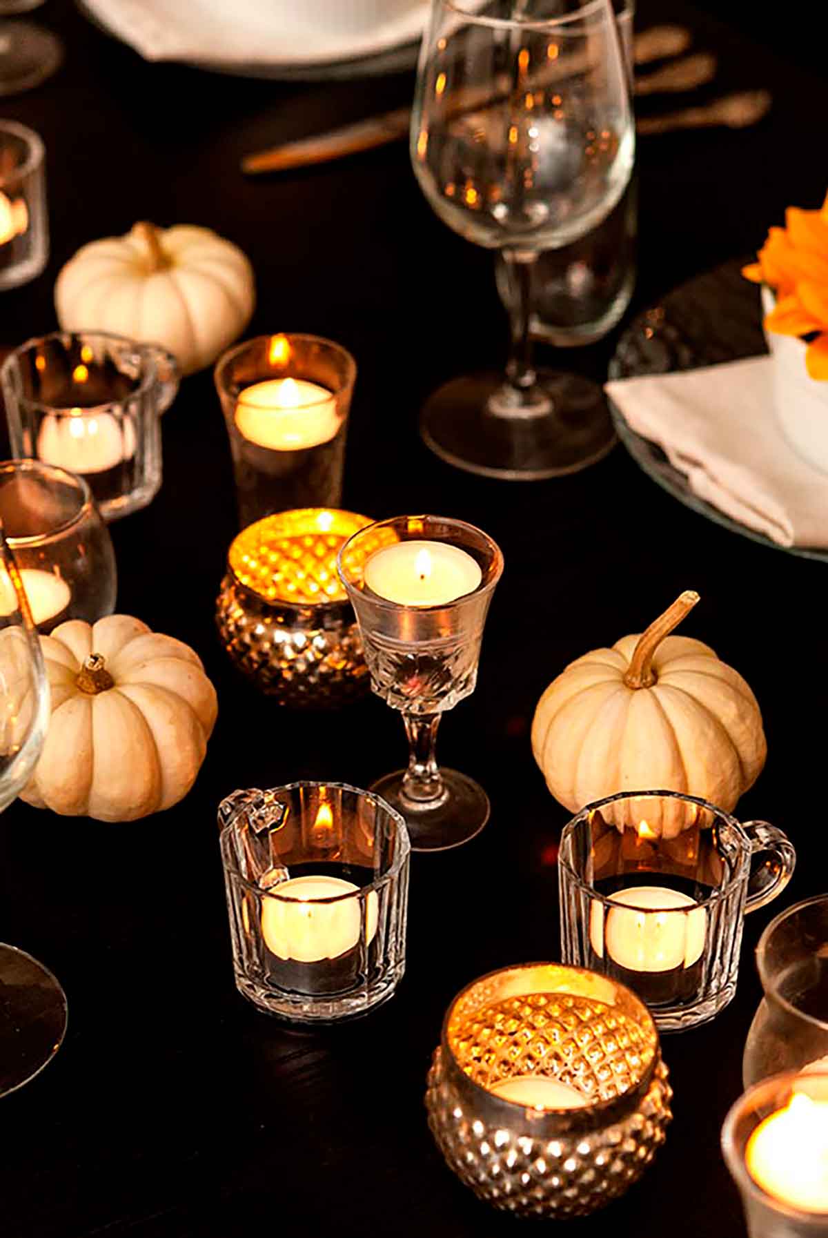 Tea lights in cordial cups and small white pumpkins on a dinner table.