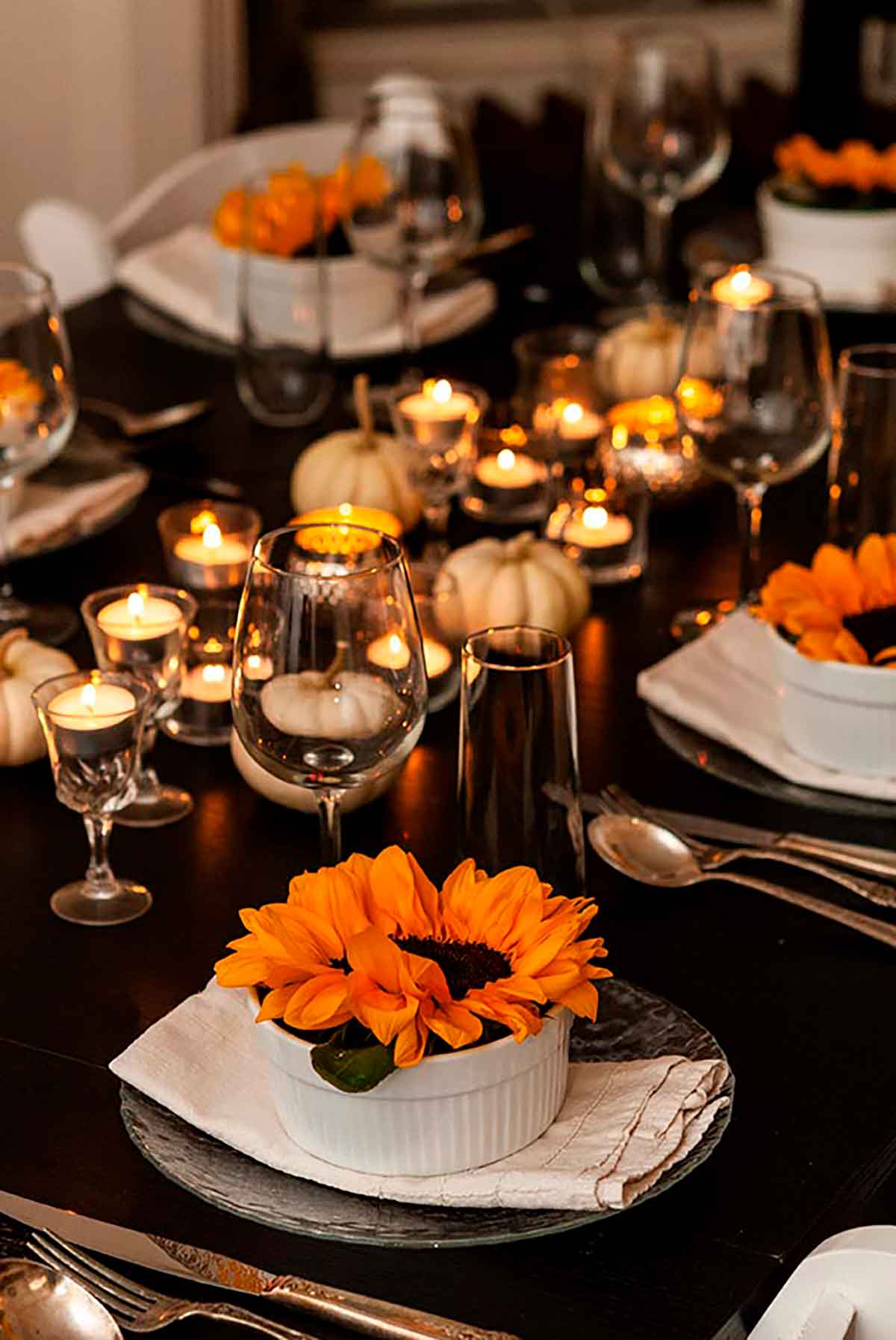 A dinner table set with tea lights, small white pumpkins, glassware and soup bowls with sunflowers inside.