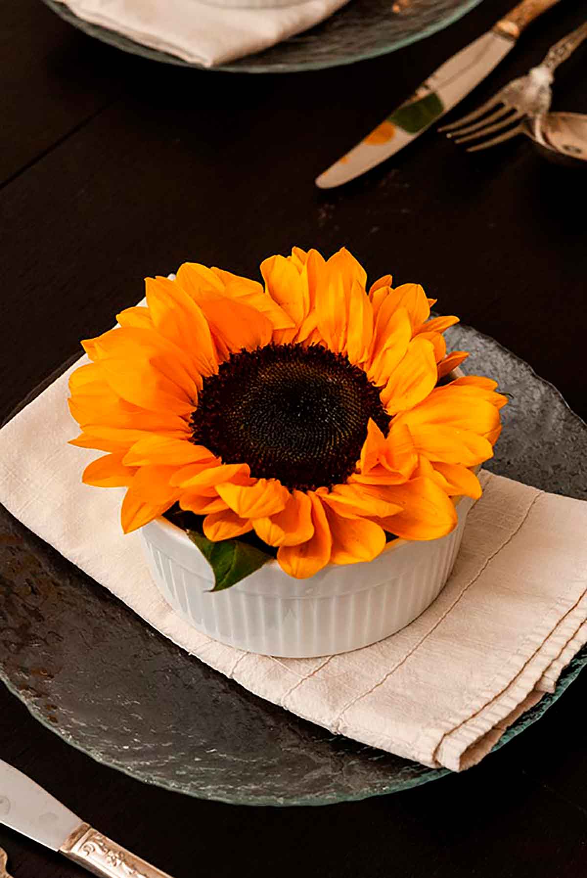 A sunflower in a soup bowl, on top of a folded napkin, on top of a plate on a table.