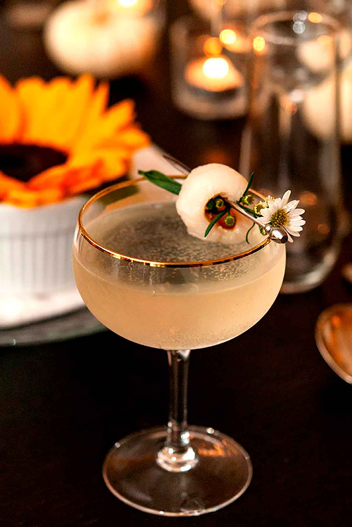 A cocktail, garnished with a lychee and small daisy, on a set dinner table with sunflowers, glasses and candles.