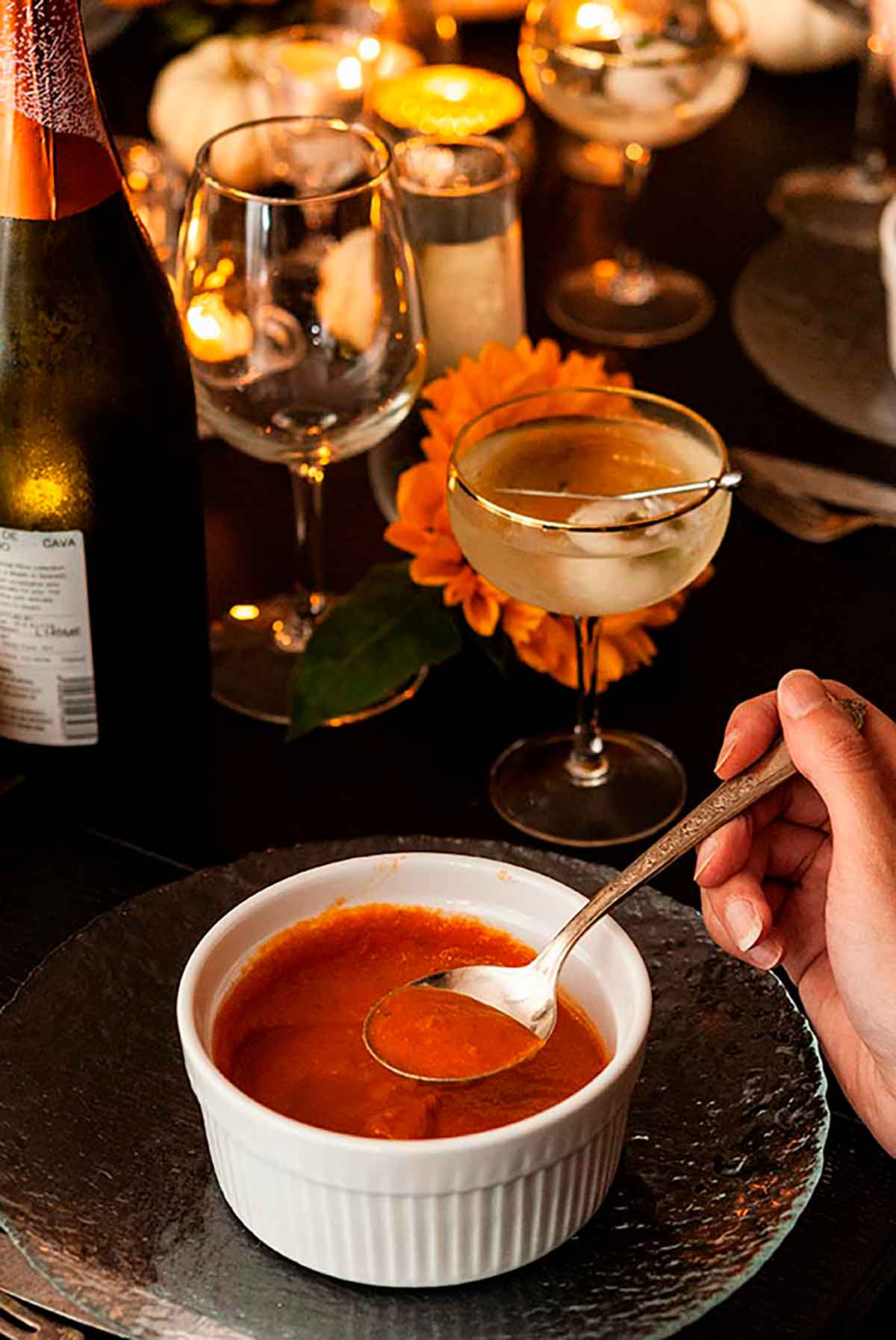 A hand holding a spoon of tomato soup over a bowl with cocktails, glassware, candles and a bottle of wine in the background.