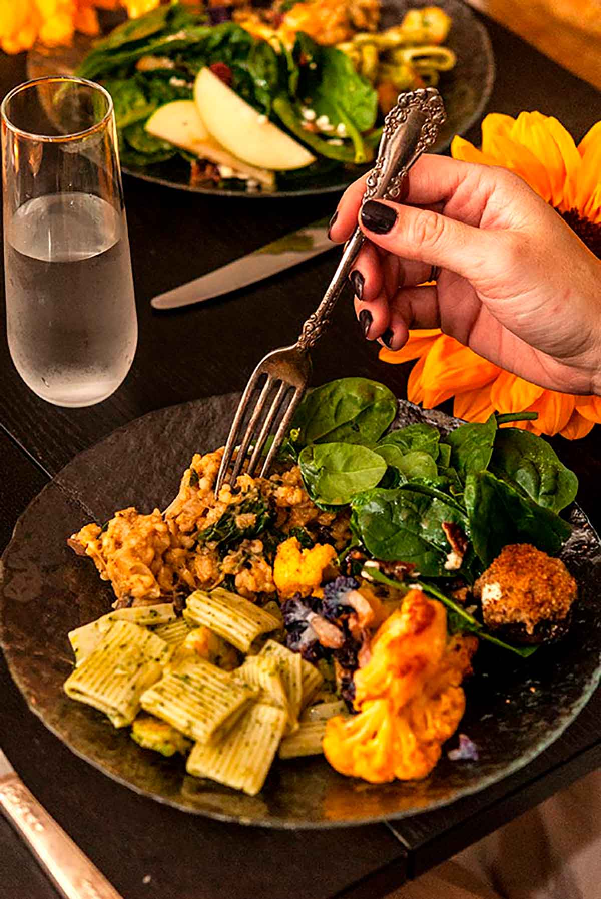 A hand holding an antique fork above a plate full of food with a sunflower and other plates of food in the background.