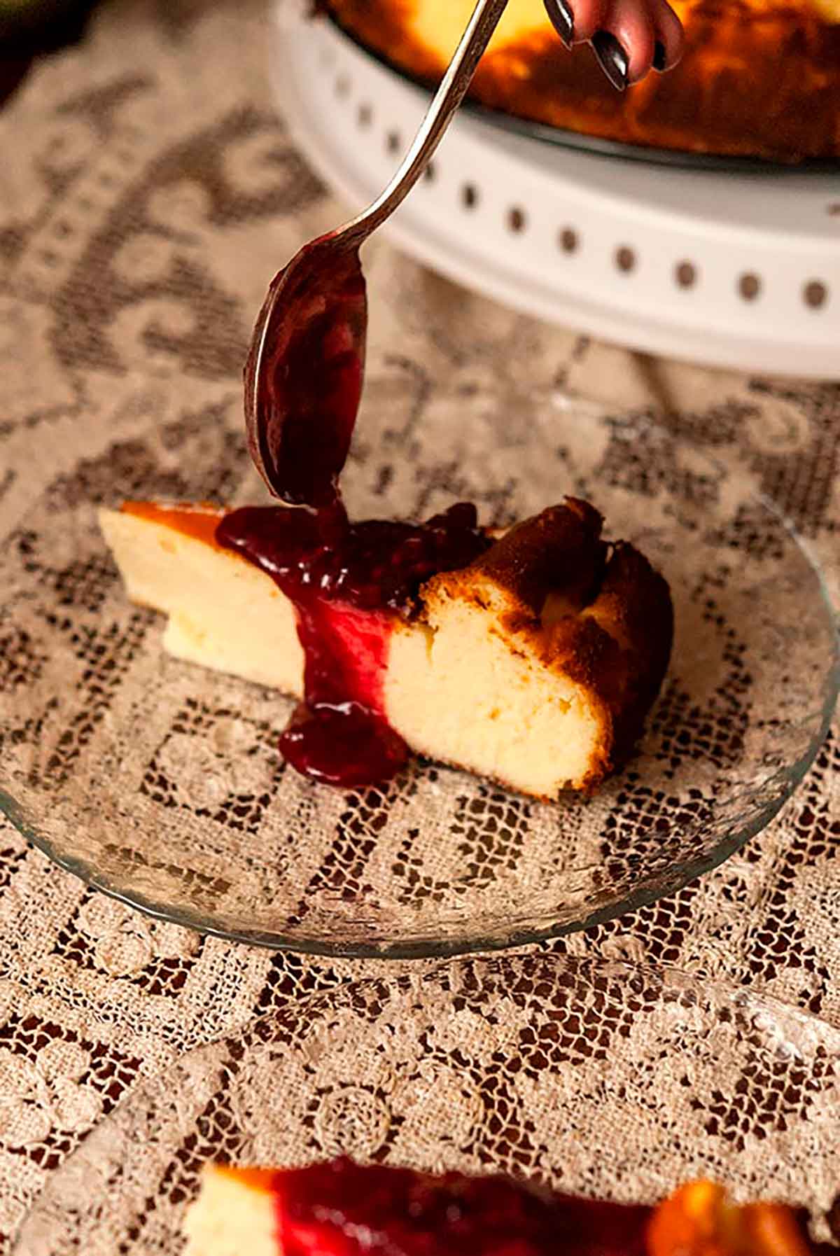 A spoon pouring raspberry sauce on a slice of cheesecake on a glass plate, on a lace tablecloth, beside a cake stand.