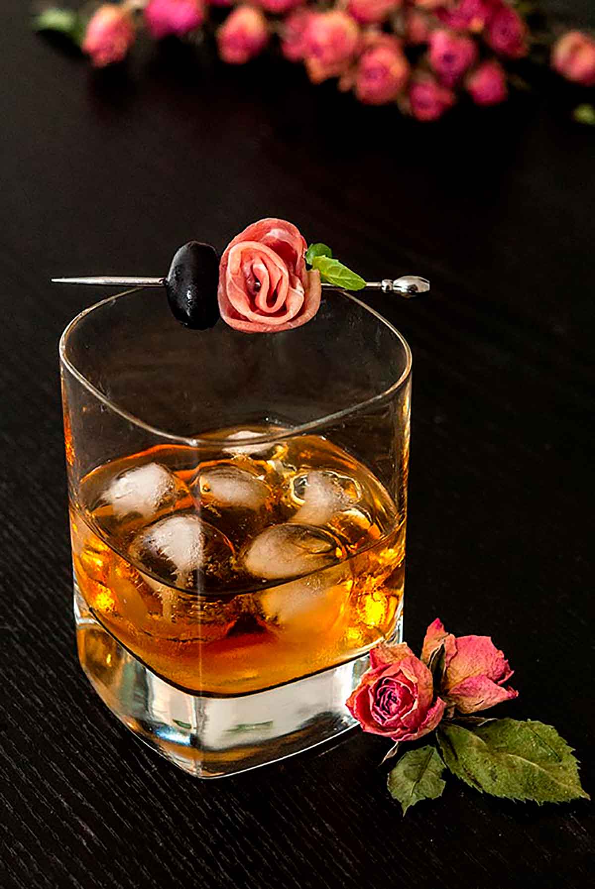 A whisky cocktail on a black table, surrounded by pink roses, garnished with a prosciutto rose and olive.