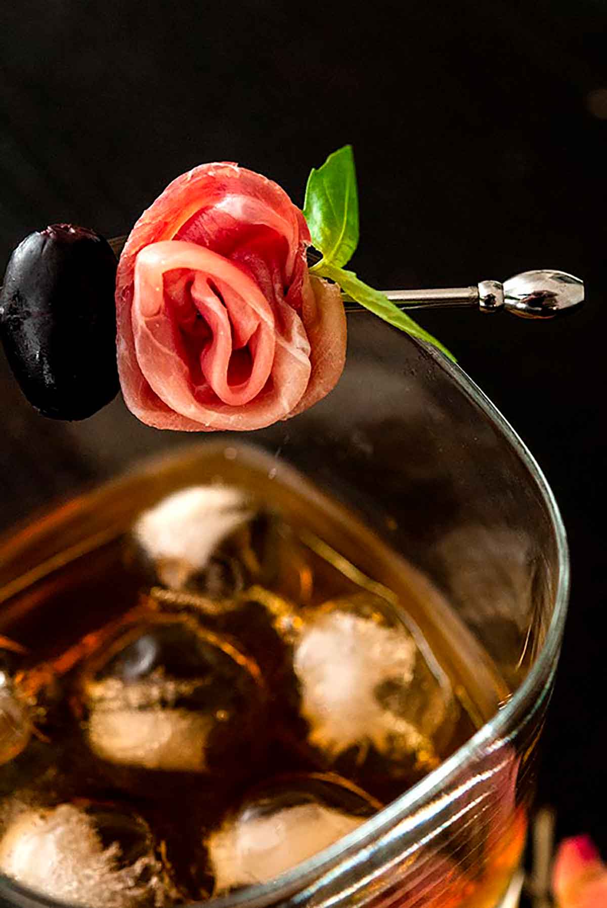 A prosciutto rose garnish on a glass filled with whisky and ice.