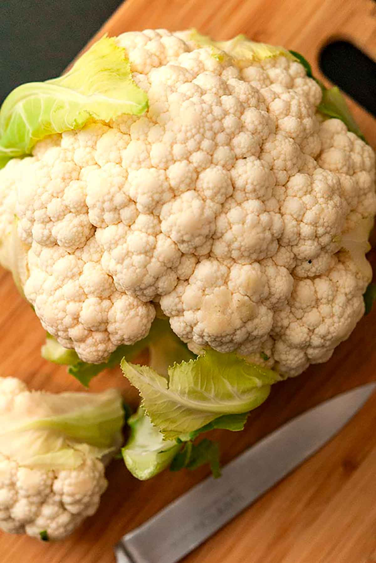 A close shot of a head of cauliflower on a cutting board beside a knife.