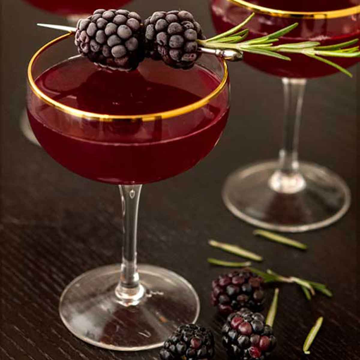 1 cocktail on a table surrounded by a few scattered blackberries, and garnished with frosted blackberries and rosemary.