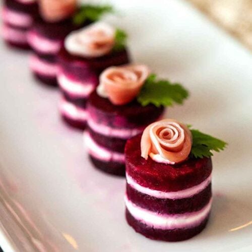 6 beet napoleons, with prosciutto roses on top, garnished with cilantro leaves on a plate.