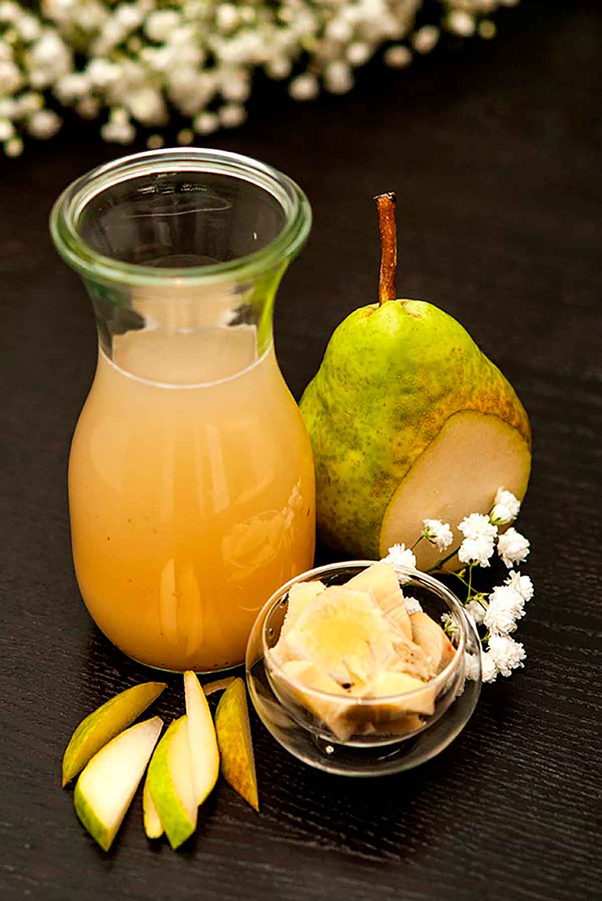 A small bottle of pear-ginger syrup on a table, along with a sliced pear, a small bowl of ginger and small pear slices.