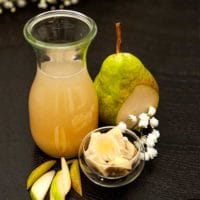 A small bottle of pear-ginger syrup on a table, along with a sliced pear, a small bowl of ginger and small pear slices.