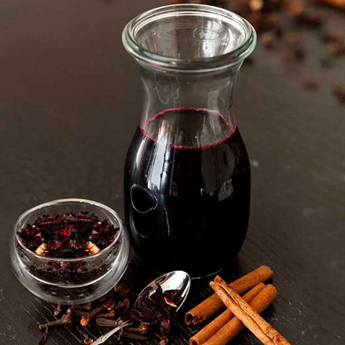 A bottle of spiced hibiscus syrup on a table surrounded by a few cinnamon sticks and a small bowl of hibiscus flowers.