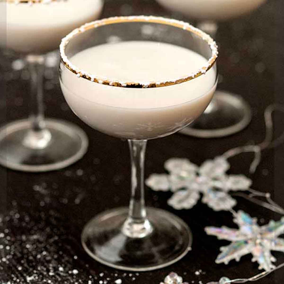 A white cocktail, rimmed with powdered sugar, on a black table, sprinkled with sugar and with a few snowflake ornaments.