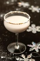 A white cocktail, rimmed with powdered sugar, on a black table, sprinkled with sugar and with a few snowflake ornaments.
