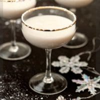 A white cocktail, rimmed with powdered sugar, on a black table, sprinkled with sugar and with a few snowflake ornaments.