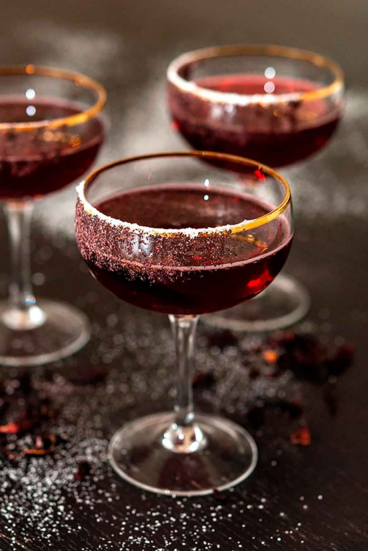 3 cocktails in coup glasses, rimmed with sugar on a sugar-sprinkled table, with a few flower petals.