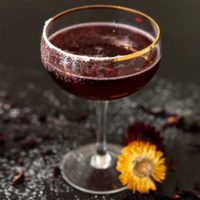 A sparkling spiced hibiscus cocktail on a dark table, sprinkled with sugar and cloves, with a tiny flower placed beside it.